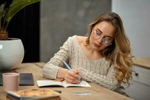 blond Modell- Blogger im Gläser, Beige Pullover. Sitzung im Küche beim hölzern Tisch, gehen zu schreiben etwas im ihr Notizbuch. schließen oben foto