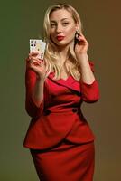 blond Dame im rot stilvoll Kleid. sie ist berühren ihr schwarz Ohrring, zeigen zwei spielen Karten, posieren auf bunt Hintergrund. Poker, Kasino. Nahansicht foto