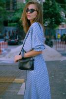 blond Mädchen im lange Blau Kleid, Sonnenbrille und ein klein schwarz Handtasche auf ihr Schulter ist Gehen allein im das Stadt. Mode und Stil. Nahansicht Schuss. foto
