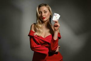 blond Modell- im rot Kleid und schwarz Ohrringe. sie ist zeigen zwei Asse, posieren auf grau Studio Hintergrund. Poker, Kasino. Nahaufnahme, Kopieren Raum foto