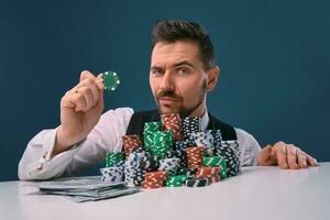 Mann im schwarz Weste und Hemd Sitzung beim Weiß Tabelle mit Stapel von Chips und Kasse auf Es, posieren auf Blau Hintergrund. Glücksspiel, Poker, Kasino. Nahansicht. foto