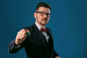 Mann im schwarz klassisch Anzug, rot Krawatte, Glasen ist zeigen zwei farbig Chips, posieren auf Blau Studio Hintergrund. Glücksspiel, Poker, Kasino. Nahansicht. foto