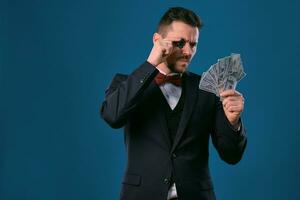 Mann im schwarz klassisch passen und rot Krawatte zeigen einer Chip und etwas Dollar Rechnungen, posieren auf Blau Hintergrund. Glücksspiel, Poker, Kasino. Nahansicht. foto