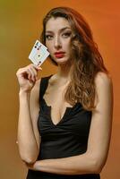 Brünette Modell- mit Ohrring im Nase, im schwarz Kleid. sie ist zeigen zwei Asse, posieren auf bunt Studio Hintergrund. Glücksspiel, Poker, Kasino. Nahansicht foto