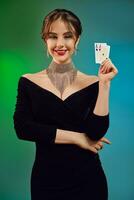 Brünette Frau im schwarz Kleid, glänzend Halskette und Ohrringe. lächelnd, zeigen zwei spielen Karten, posieren auf bunt Hintergrund. Poker, Kasino. Nahansicht foto