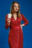 blond Modell- mit bilden, tätowiert Hand, im rot Paillette Kleid ist lächelnd, zeigen zwei Asse, posieren auf Blau Hintergrund. Poker, Kasino. Nahansicht. foto