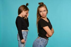 Mama und Tochter mit ein komisch Frisuren, gekleidet im schwarz Hemden und Blau Denim Jeans sind posieren gegen ein Blau Studio Hintergrund. Nahansicht Schuss. foto