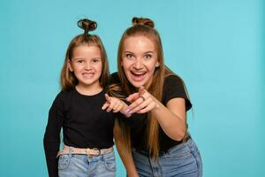 Mama und Tochter mit ein komisch Frisuren, gekleidet im schwarz Hemden und Blau Denim Jeans sind posieren gegen ein Blau Studio Hintergrund. Nahansicht Schuss. foto