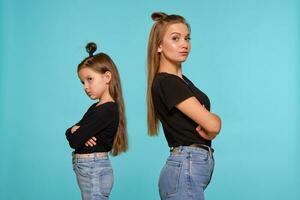 Mama und Tochter mit ein komisch Frisuren, gekleidet im schwarz Hemden und Blau Denim Jeans sind posieren gegen ein Blau Studio Hintergrund. Nahansicht Schuss. foto