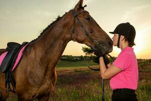 schön lächelnd Mädchen Jockey Stand Nächster zu ihr braun Pferd tragen Besondere Uniform auf ein Himmel und Grün Feld Hintergrund auf ein Sonnenuntergang. foto