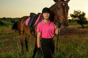 schön lächelnd Mädchen Jockey Stand Nächster zu ihr braun Pferd tragen Besondere Uniform auf ein Himmel und Grün Feld Hintergrund auf ein Sonnenuntergang. foto