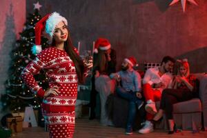 Porträt von ein jung Frau mit ein Glas von Champagner beim Zuhause auf das Vordergrund. schön Brünette im ein Santa Hut, rot Kostüm mit Hirsche foto