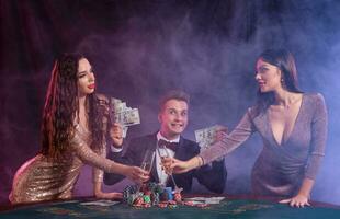 Mann spielen Poker beim Kasino Sitzung beim Tabelle mit Stapel von Chips, Geld, Karten. feiern Sieg mit zwei Frauen. Schwarz, Rauch Hintergrund. Nahansicht. foto