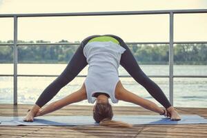 Frau im Weiß Unterhemd und schwarz Gamaschen ist üben Yoga durchführen Yoga-Asanas auf ein Matte draußen, in der Nähe von ein Flussufer. foto