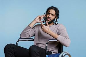 verwirrt arabisch Mann mit physisch Beeinträchtigung antworten Smartphone Anruf während Sitzung im Rollstuhl und suchen beim Kamera. Person mit Behinderung Sprechen auf Handy, Mobiltelefon Telefon Porträt foto
