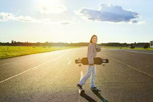 jung Skater Mädchen, Teenager Skaten auf Kreuzer, halten Longboard und Gehen auf Beton leeren Straße foto