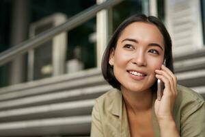 Porträt von asiatisch Mädchen lächelt während Gespräche auf Handy, Mobiltelefon Telefon. jung Frau Berufung ein Freund, Sitzung auf Treppe foto