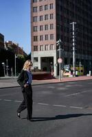Vertikale Porträt von zuversichtlich und stilvoll Geschäftsfrau im Anzug, gehen, Kreuz das Straße im Stadt Center, trägt Sonnenbrille foto