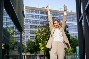 Porträt von glücklich jung Geschäftsfrau Aufzug Hände hoch, tut Faust Pumpe, feiert Sieg, Gewinnt etw, erreicht Ziel, steht draußen auf Straße foto