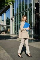 Porträt von Geschäftsfrau im Beige passen und hoch Absätze Gehen auf Straße. korporativ Frau gehen zu arbeiten, halten Mappe mit Unterlagen foto