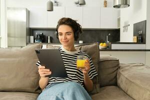 Porträt von Frau im Kopfhörer, Sitzung auf Sofa mit Tablette und Trinken Orange Saft, Aufpassen Fernseher Show auf ihr Gerät, mit Sozial Medien Anwendung, Ausgaben Zeit beim Zuhause foto