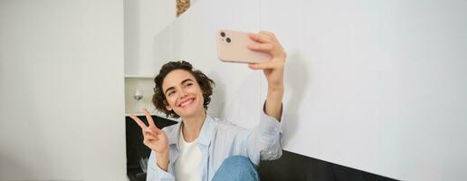 Porträt von schön Brünette Mädchen, nimmt Selfie auf Smartphone im ihr Küche, posieren zum Foto auf Handy, Mobiltelefon Telefon