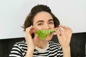 schließen oben Porträt von jung Frau, Vegetarier Mädchen, Likes Essen Gemüse, posieren mit Grüner Salat Blatt und lächelnd, posieren im das Küche. Konzept von gesund Essen und Diät foto