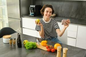 Porträt von schön jung Frau, Essen Blogger Aufzeichnung Video von ihr Kochen im das Küche, zeigen Mahlzeit Zutaten, vorbereiten ein Mahlzeit auf Kamera, mit Smartphone zum Leben Streaming foto