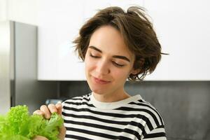 schließen oben Porträt von lächelnd, schön weiblich Modell, Herstellung ein Salat, halten Grün Grüner Salat Blatt, vorbereiten Gemüse, Kochen im das Küche, Stehen im gestreift T-Shirt foto