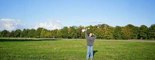 aufgeregt jung Frau Aufzug ihr Ukulele oben Über Kopf und lächelt, posieren mit Musical Instrument im Grün Feld Park foto