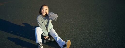 glücklich schön Koreanisch Teen Mädchen sitzt auf ihr Skateboard, Kreuzfahrt auf Longboard, tragen beiläufig Kleider foto