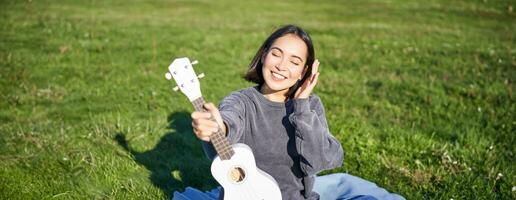 lächelnd asiatisch Mädchen mit Ukulele, spielen im Park und Singen, Lebensstil Konzept foto