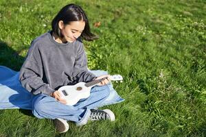 lächelnd asiatisch Mädchen, Musiker spielen Ukulele im Park, suchen mit Pflege und Liebe beim ihr Instrument, Sitzung auf Decke auf sonnig Tag foto