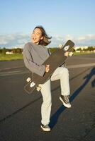 komisch asiatisch Mädchen genießen skaten, halten Skateboard mögen Gitarre und Schatten spielen, haben Spaß draußen foto