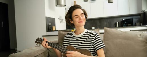 schließen oben Porträt von schön lächelnd Frau, spielen Ukulele, klimpern Saiten mit geschlossen Augen und zufrieden Gesicht, Sitzung im Leben Zimmer foto