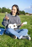 Vertikale Schuss von lächelnd asiatisch Mädchen mit Smartphone, spielen Ukulele, lesen Akkorde oder Text während Singen, entspannend draußen. Lebensstil und Menschen Konzept foto