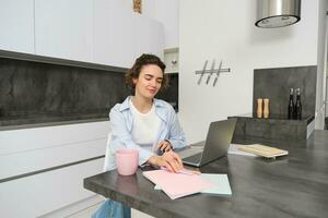 Porträt von Brünette Frau tut Hausaufgaben, funktioniert von heim, bereitet vor zum Prüfung im Küche, sitzt mit Notizbuch und Laptop foto