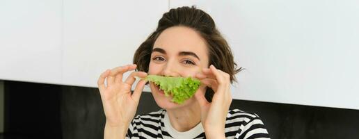 schließen oben Porträt von jung Frau, Vegetarier Mädchen, Likes Essen Gemüse, posieren mit Grüner Salat Blatt und lächelnd, posieren im das Küche. Konzept von gesund Essen und Diät foto