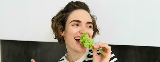 Porträt von sorglos Vegetarier Mädchen, Essen Gemüse, beißen Grüner Salat Blatt mit glücklich lächelnd Gesicht, haben gesund Snack, Likes Gemüse, steht im das Küche foto