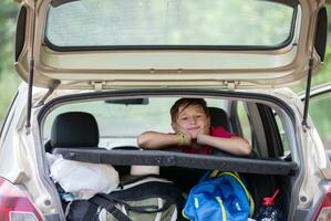 ein Kind auf ein Reise. ein Junge sieht aus aus von das Kofferraum von ein Auto gefüllt mit enorm Taschen. foto