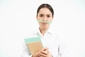 komisch Koreanisch Frau mit Notizbücher, hält Stift mit Lippen und Nase und macht Gesichter, steht Über Weiß Studio Hintergrund foto