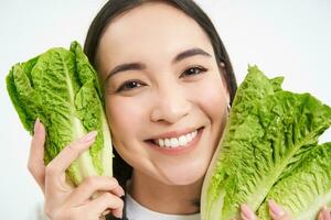 schließen oben Porträt von glücklich Koreanisch Frau, zeigt an ihr Gesicht mit Kopfsalat, Essen Kohl, Likes Gemüse, fühlt sich gesund und energiegeladen, Weiß Hintergrund foto