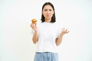 verärgert asiatisch Frau, halten Cupcake und beschwert sich, will zu Essen Müll Süss Essen, steht Über Weiß Hintergrund foto