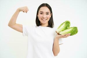 stark asiatisch Frau zeigt an frisch Gemüse, Grüner Salat und ihr Muskeln, Biegen Bizeps mit lächelnd Gesicht, Weiß Hintergrund foto