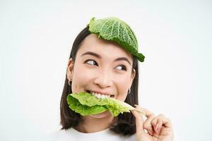schön asiatisch Mädchen mit Grüner Salat auf Kopf, lächelnd und Essen Kohl Blatt, vegan mit Gemüse, Weiß Hintergrund foto