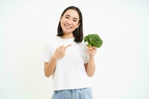 Porträt von süß asiatisch Frau, zeigen beim Brokkoli, zeigen Gemüse, Ernährungsberater empfehlen gesund Diät Essen, Weiß Hintergrund foto
