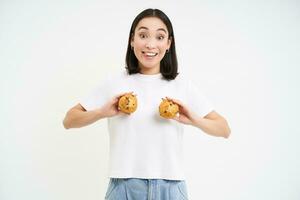 glücklich jung Koreanisch Frau, zeigen zwei Cupcakes Über Brust, lächelnd und suchen Glücklich, Gebäck und Bäckerei Konzept foto