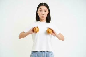 glücklich japanisch Frau zeigt an Cupcakes auf Brust, Essen Gebäck, Stehen Über Weiß Hintergrund foto