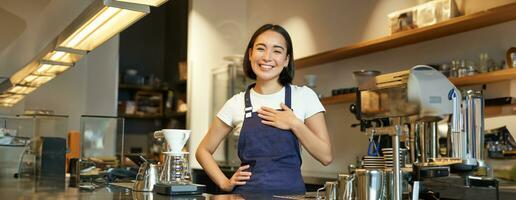 freundlich lächelnd asiatisch Mädchen Barista, hält Hände auf Herz, sieht aus dankbar beim Klient, liebt ihr Gäste im Cafe, steht im Uniform Schürze foto