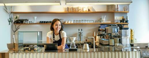 lächelnd freundlich asiatisch Mädchen Barista, tragen Schürze, Arbeiten beim Schalter, brauen Filter Kaffee, Verwaltung Cafe Geschäft foto